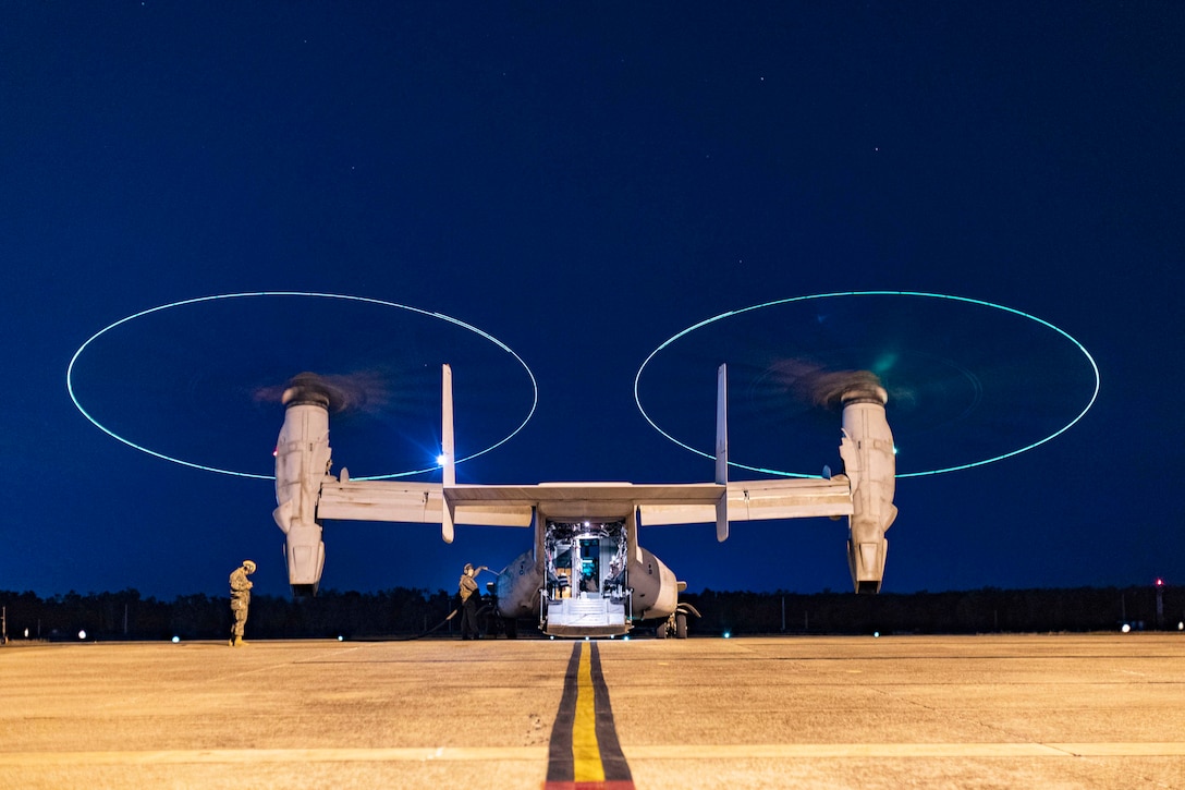 An aircraft with an open doorway sits on a tarmac in the dark illuminated by blueish lights as Marines work nearby.