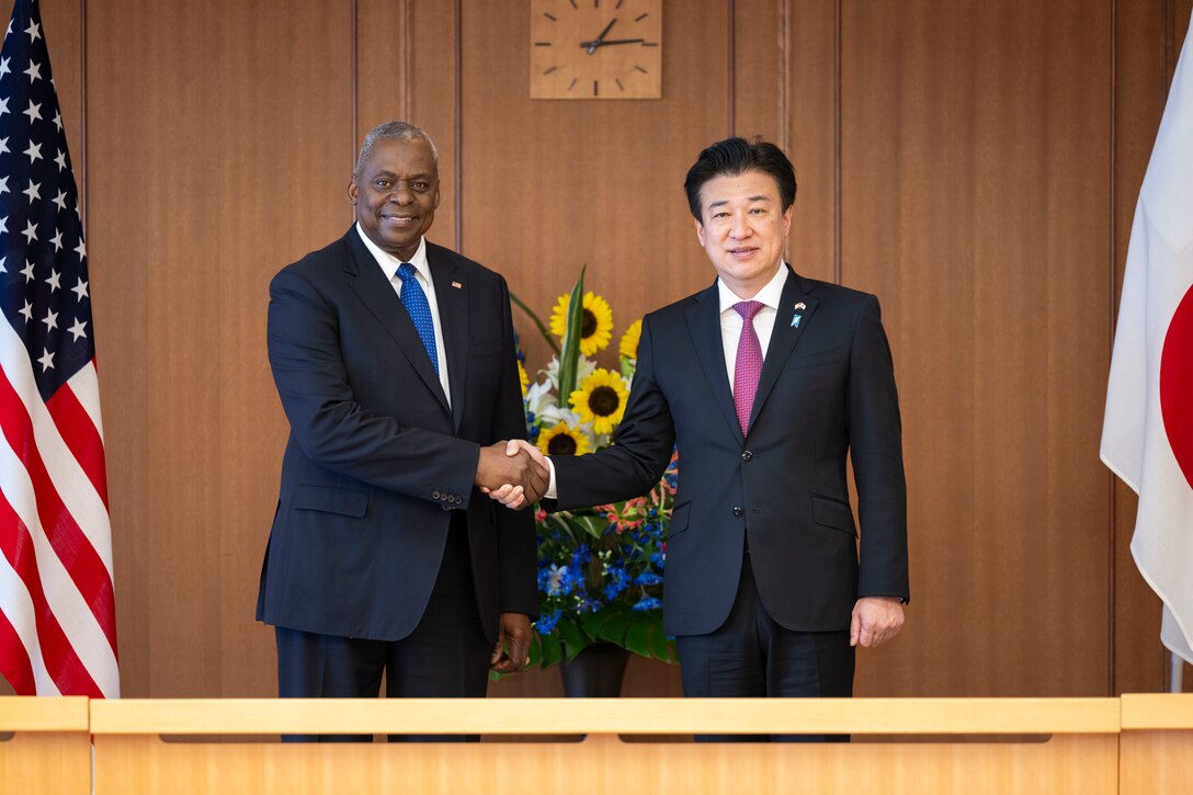 U.S. Secretary of Defense Lloyd J. Austin III shakes hands with a Japanese official, both smiling, in a formal setting with flags and flowers.