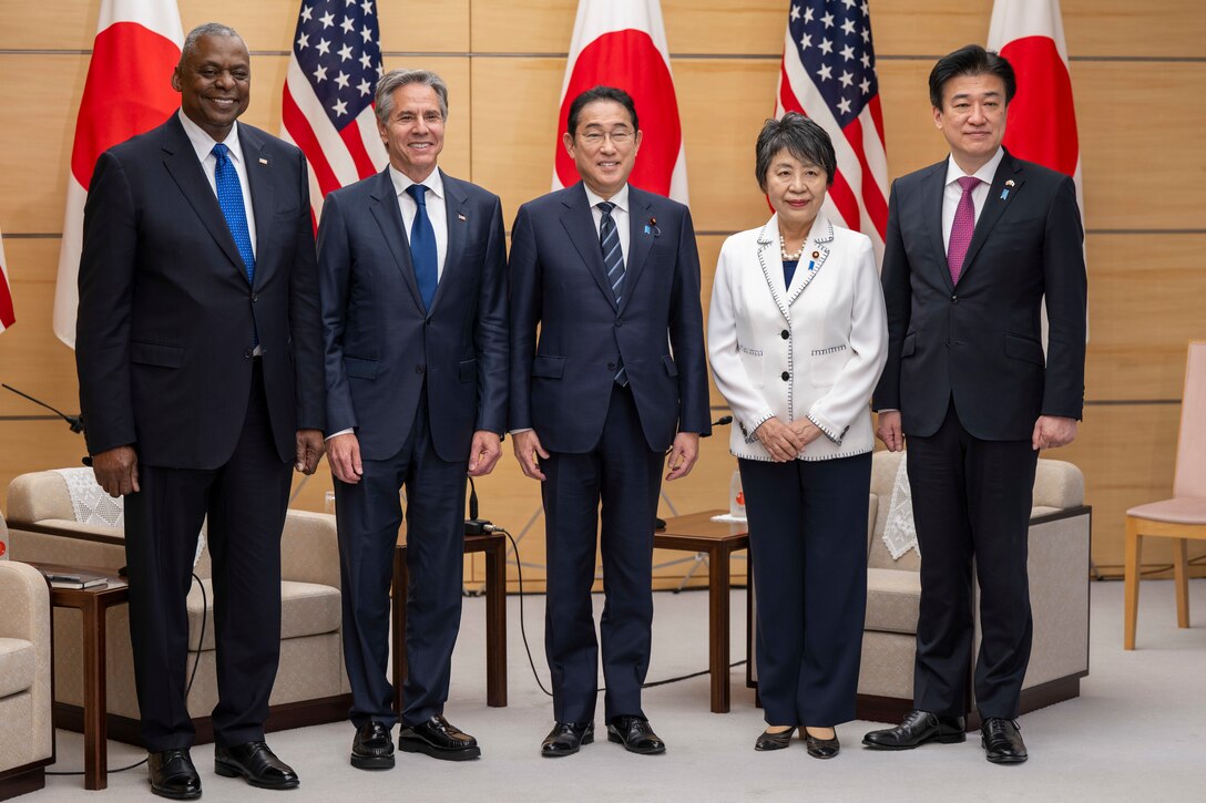 Five officials pose together, with flags of Japan and the U.S. in the background, in a formal setting.