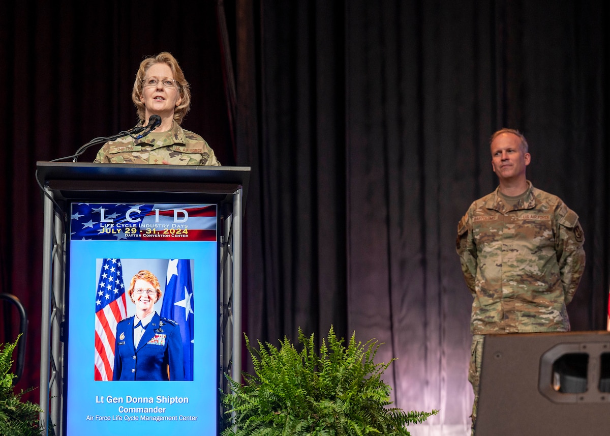 Woman stands behind podium. Man stands nearby. Both in USAF uniforms.
