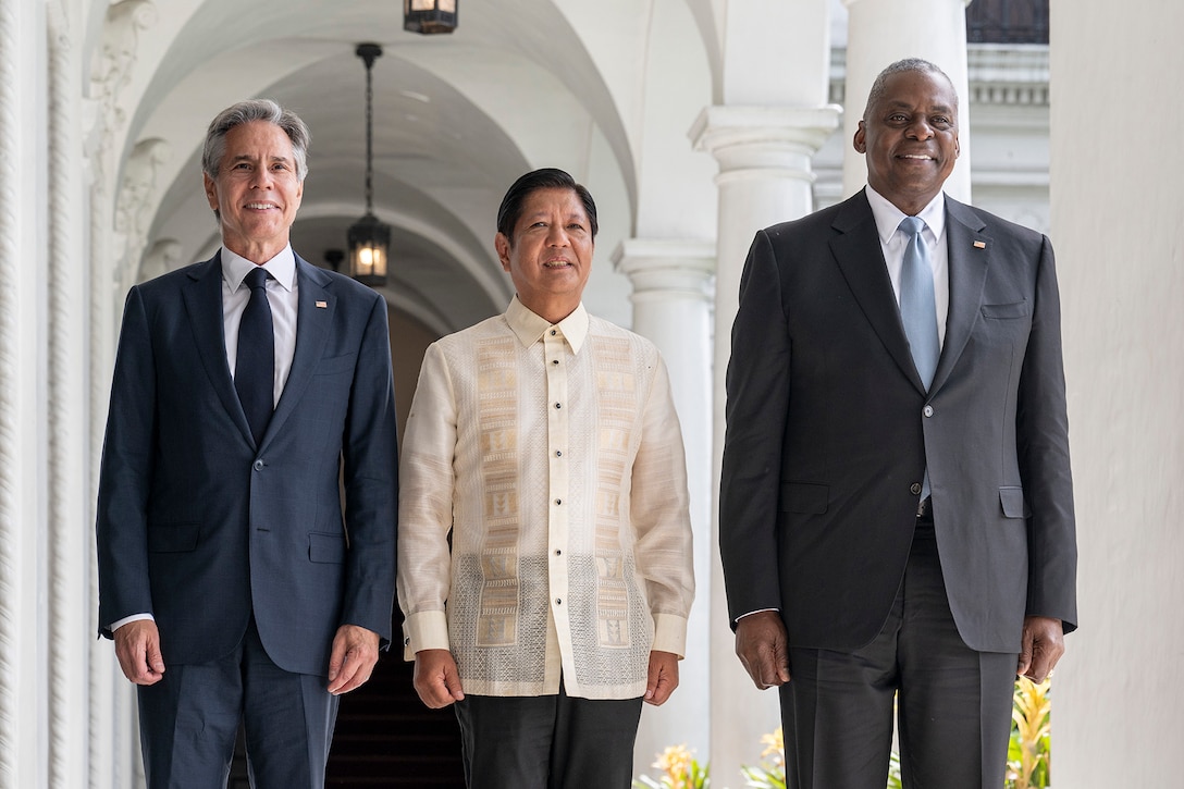 Three civilians stand and smile for a photo outdoors by columns of an ornate white building.