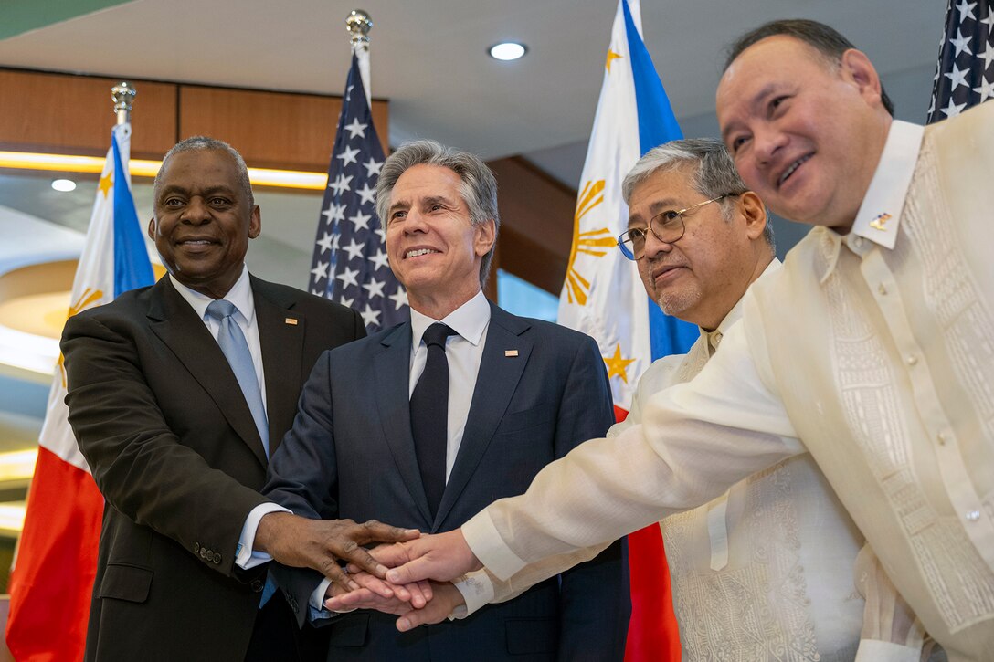Four civilian officials stand and smile while joining their hands together in front of them.