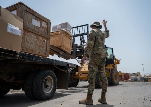 A uniformed servicemember gives hand signals to another servicemember driving a forklift, which is carrying two crates
