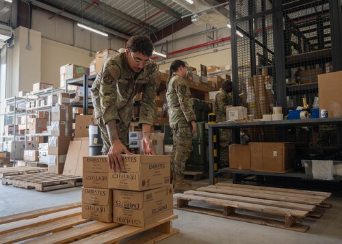 A uniformed servicemember picks up a box in a warehouse. In the background are 2 more servicemembers stocking a shelf