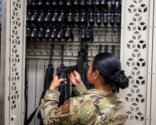 A female uniformed servicemember inspects a handgun in front of a locker stocked with more weapons