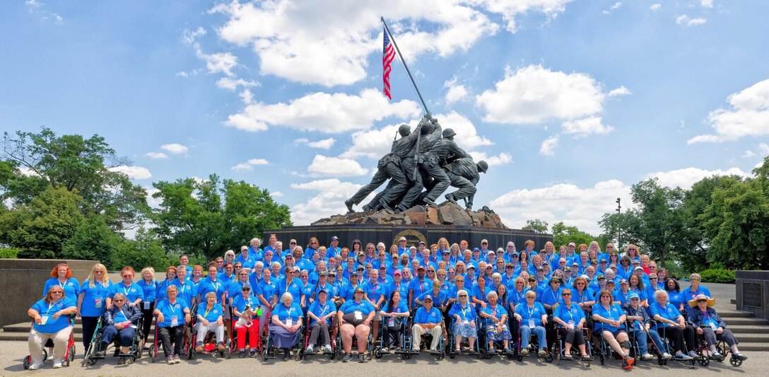 150 female Veterans in blue shirts pose for an group photo in front of the US Marine Corps War Memorial with blue sky's and light clouds. the memorial shows Marines raising the US flag over Iwo Jima.