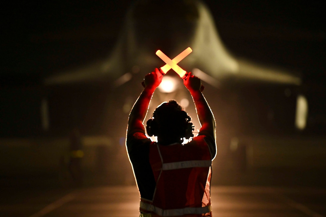 An airman uses light sticks to guide a large aircraft on a runway at night.