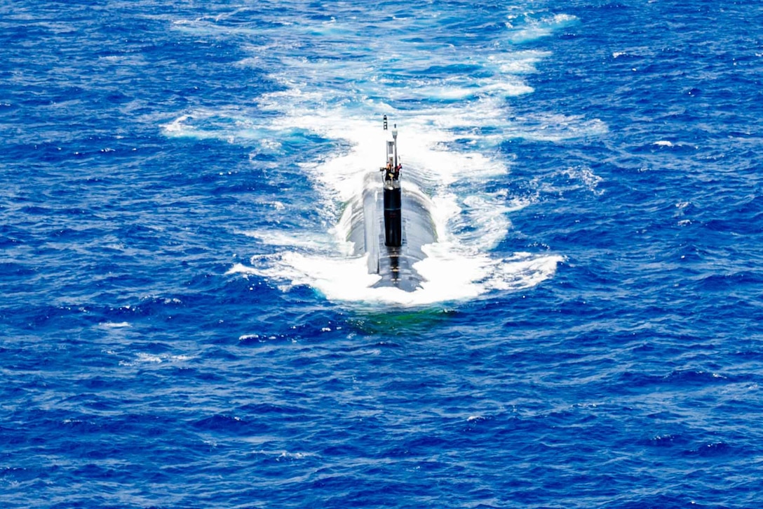 The top of a submarine is seen as it steams through blue water.