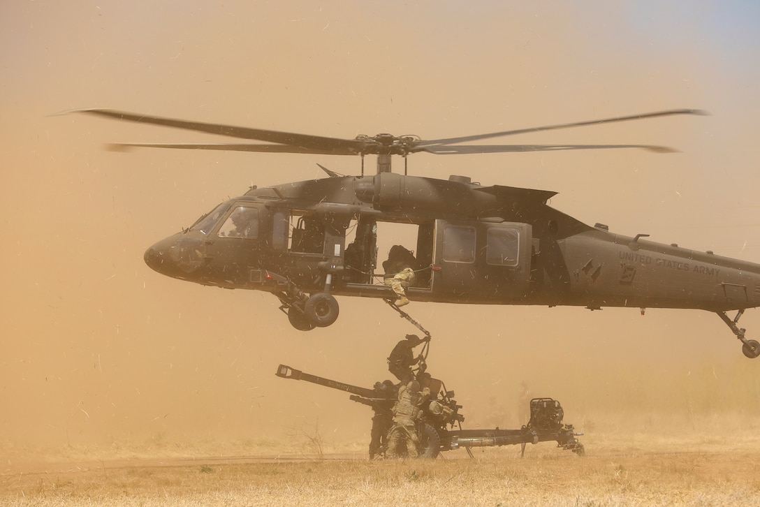 Soldiers attach a military weapon to a helicopter hovering over them creating a plume of orange dust.