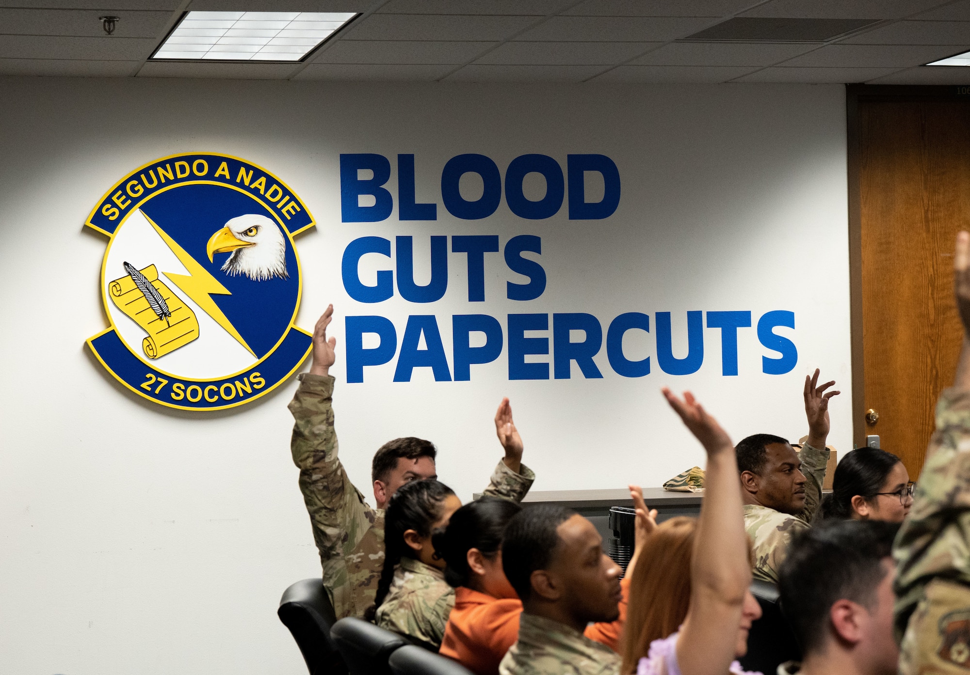 A group of people in business casual and military attire sit in a conference room raising their hands during a meeting. A sign reading "Bloods, Guts, and Papercuts" is on the wall behind them.