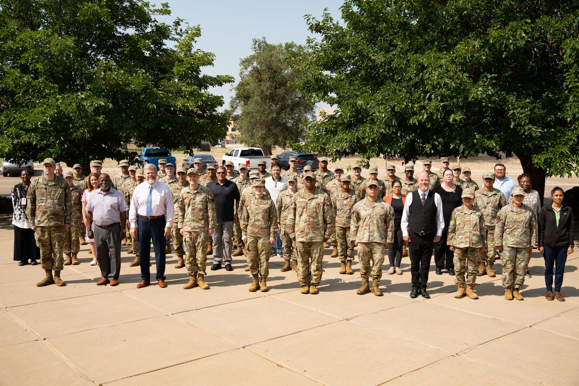 A group of military members and civilians stand in formation for a group photograph outside.