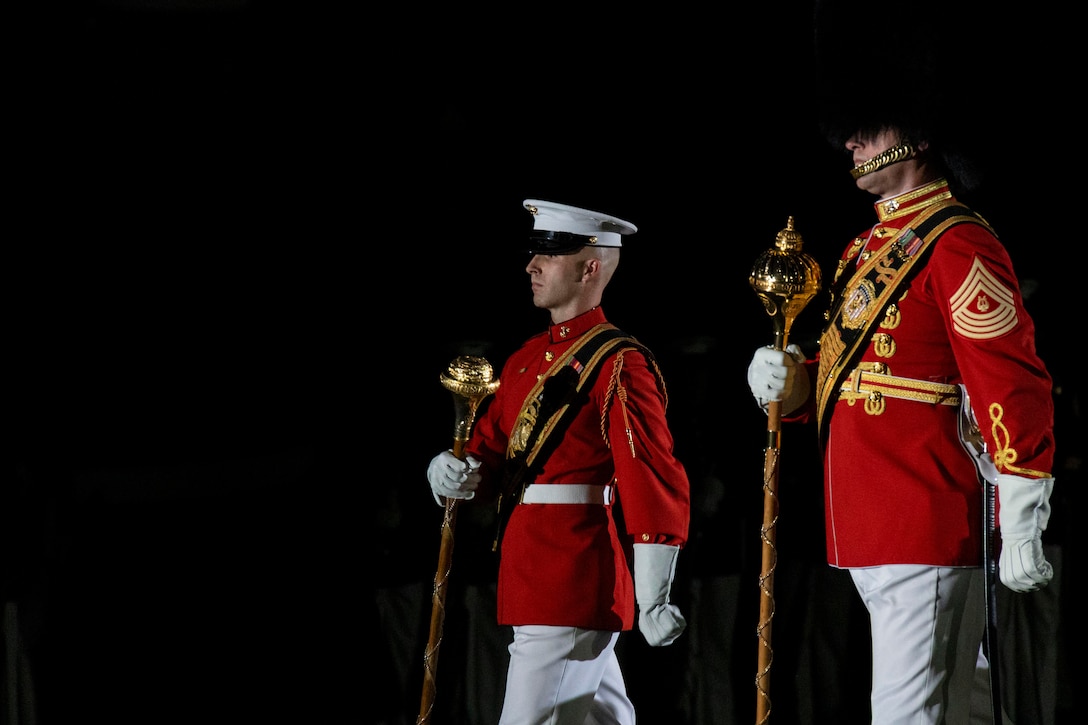 Two Marines in ceremonial dress walk in formation while carrying golden sticks in the dark.