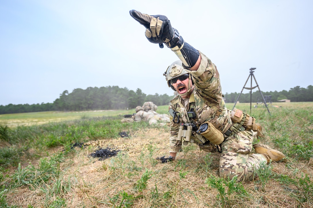 A soldier shouts while laying in a field and pointing their finger as fellow soldiers aim a weapon background.