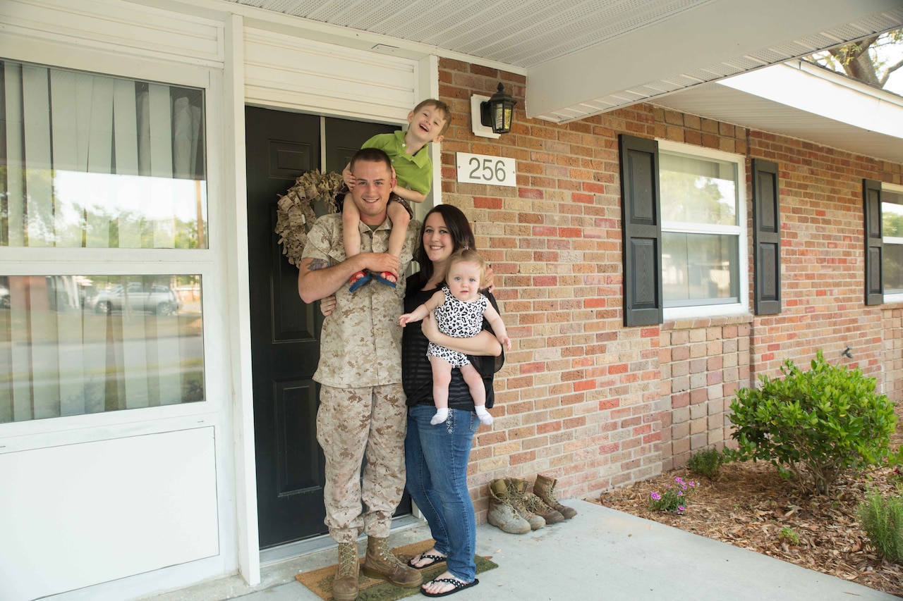 A child sits on the shoulder of a service member in uniform on a porch next to a woman in civilian attire holding another child.