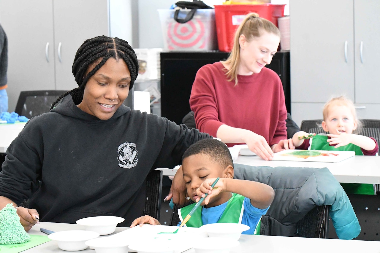 A woman sits at a table next to a child holding a paint brush.