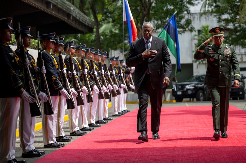 A person in business attire places their hand on their chest as a person in a foreign military uniform salutes while an honor cordon stands at attention.