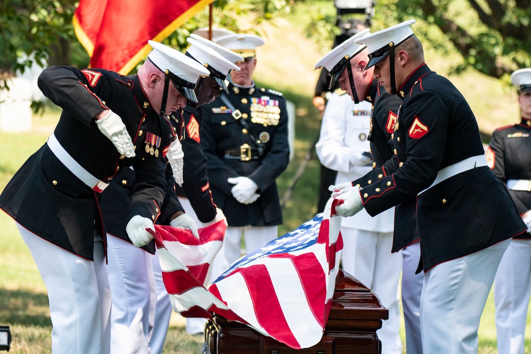 A group of Marines in dress uniform stand on either side of a casket and drape it with the American flag.