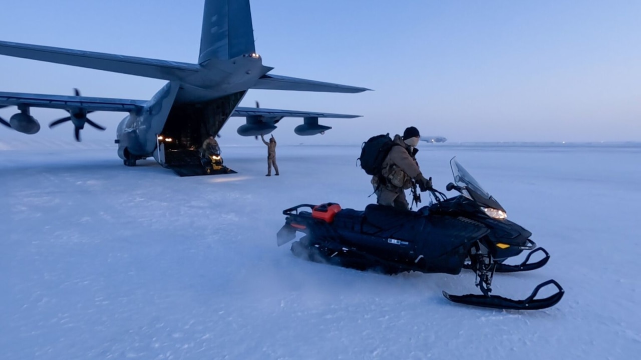 A uniformed service member starts a snowmobile near the back of a plane with an open cargo bay.