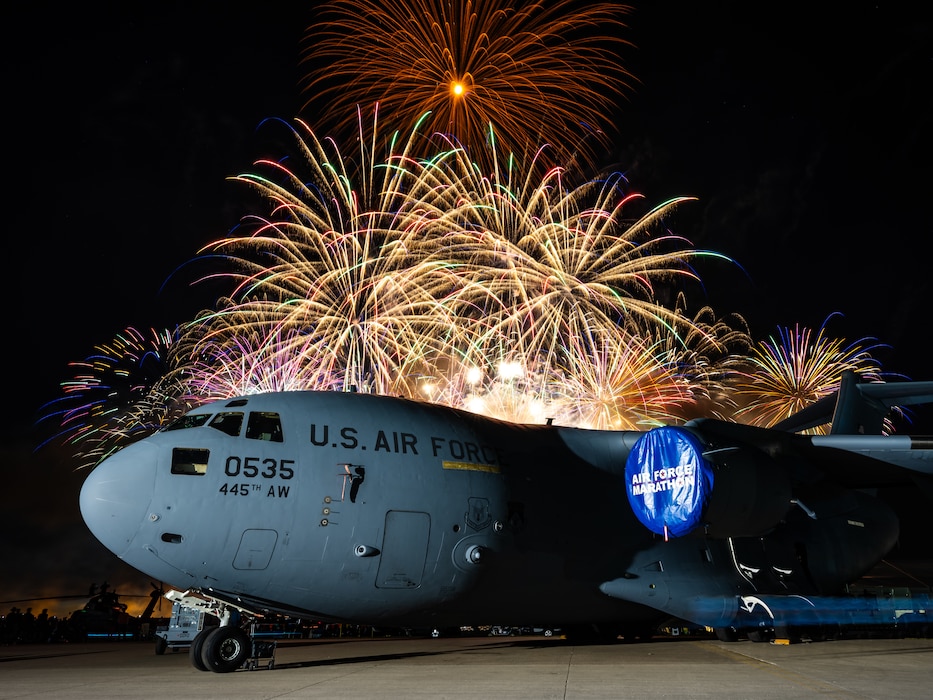 A C-17 aircraft is illuminated in the foreground with bright colorful firework display over top in the background