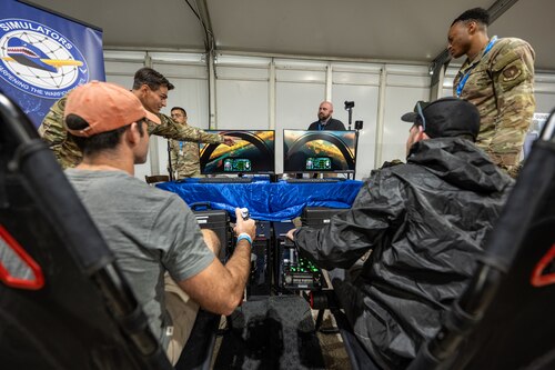 Two airshow attendees sit with backs to camera in foreground looking at two fighter jet simulation monitors with two uniformed members supervising