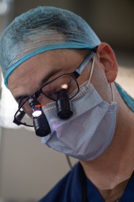 A health provider in sterile clothing examines the teeth of patient using a magnifier on his glasses.
