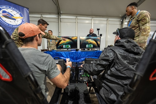 Two airshow attendees sit with backs to camera in foreground looking at two fighter jet simulation monitors with two uniformed members supervising