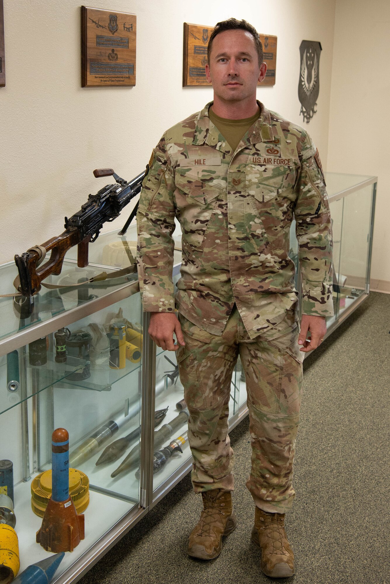 A man in military uniform poses for a photo in front of a variety of military weapons and ordnance on display.