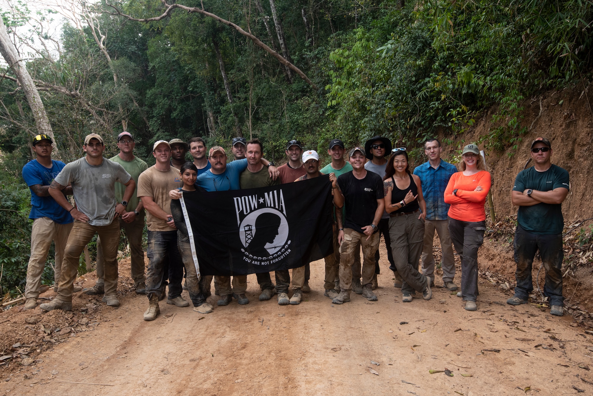 A group of men and women smile posing for a picture with a jungle in the background, while holding a flag that says POW/MIA "You are not forgotten."