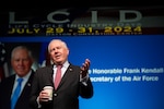 Secretary of the Air Force talks on stage. // Man in suit holding coffee gives speech in front of blue background.