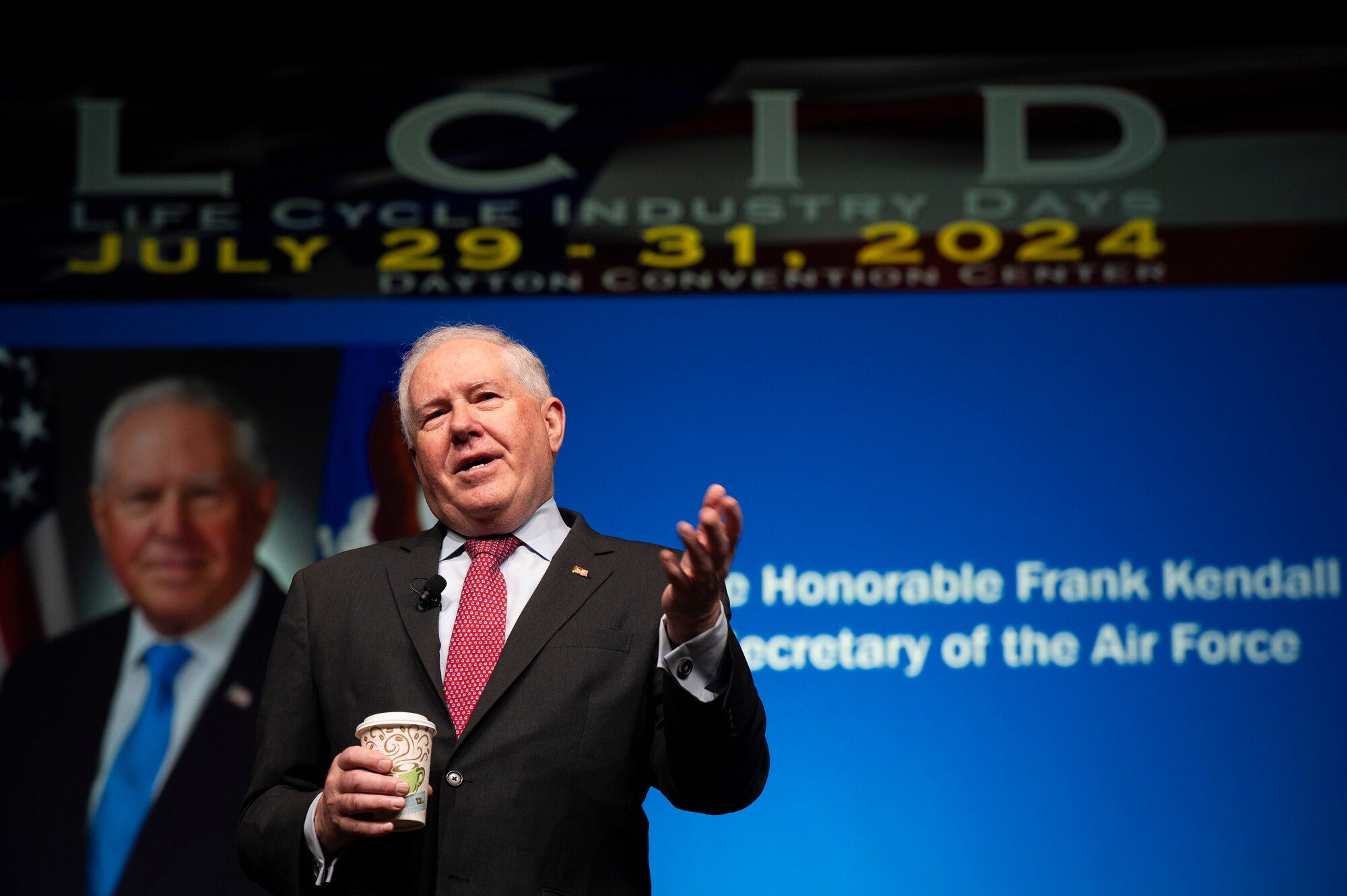 Secretary of the Air Force talks on stage. // Man in suit holding coffee gives speech in front of blue background.
