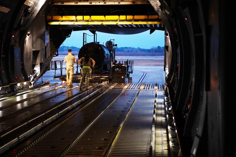 Two airmen watch as heavy equipment is loaded onto the loading bay of a large aircraft.