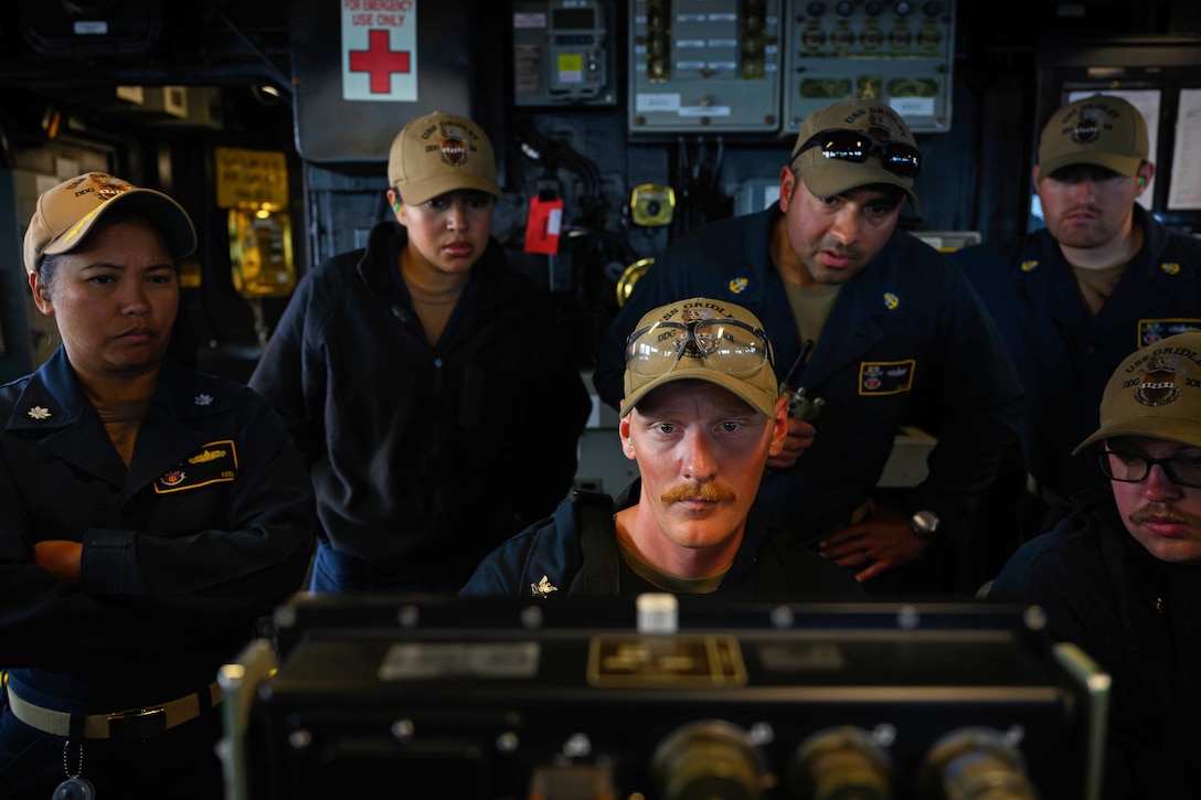 Six sailors look intently at a weapons system monitor in a darkened room.