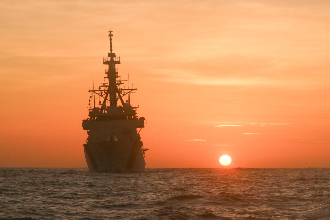 A large ship floats on the water as the sun sits low in an orange sky.