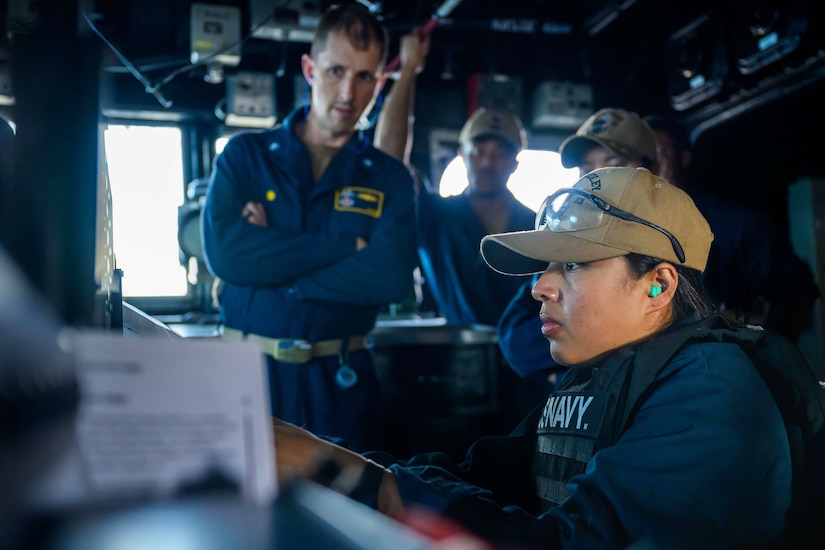 A sailor looks intently at a weapons system monitor on the bridge of a ship as other sailors watch.