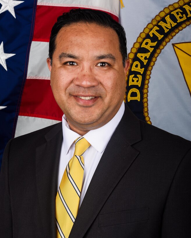 A man sits for an official portrait in front of the American flag and the Senior Executive Service flag