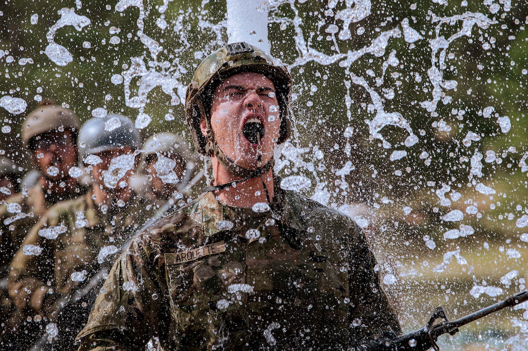 A uniformed Air Force cadet holding a weapon shouts as water falls. Other cadets are visible in the background.