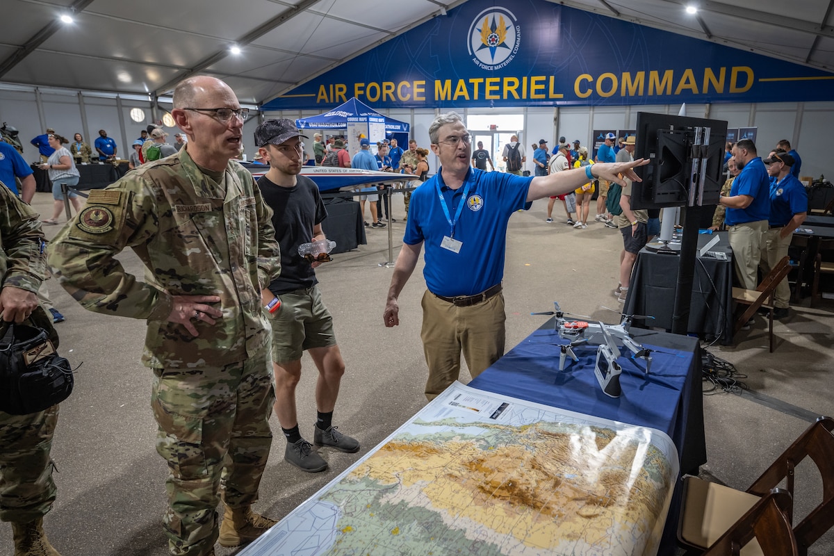 Gen. Richardson stands with hands on hip and shocked expression as he is briefed by an AFMC personnel in blue polo