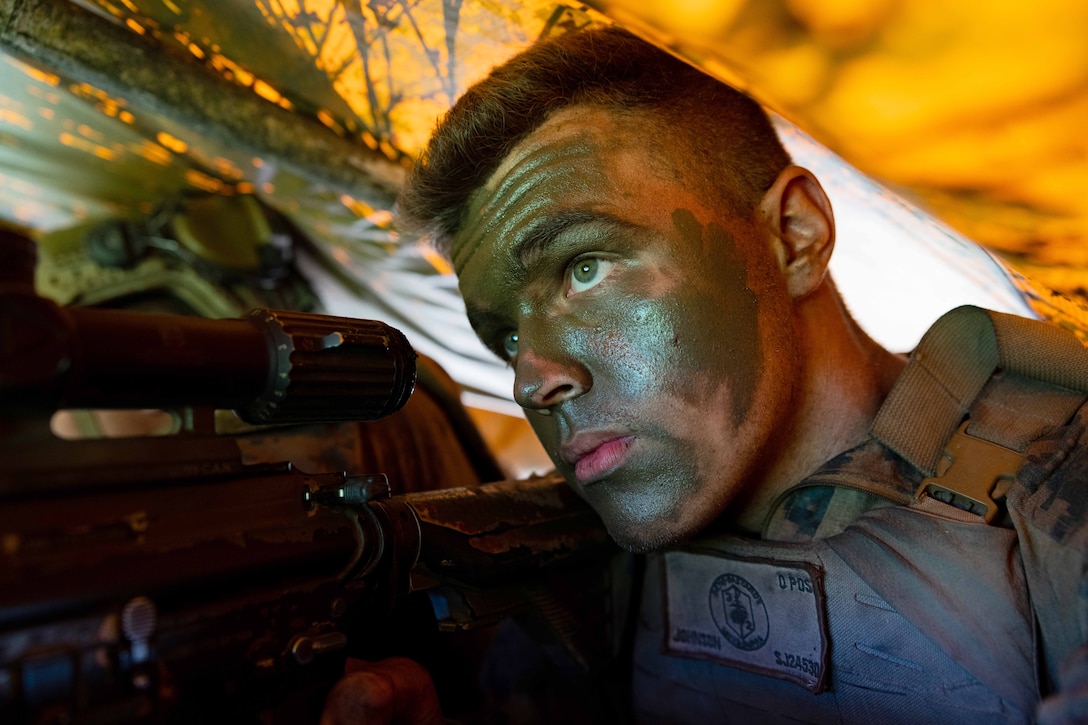 A close-up of a Marine wearing camouflage aiming a weapon out of a tent.