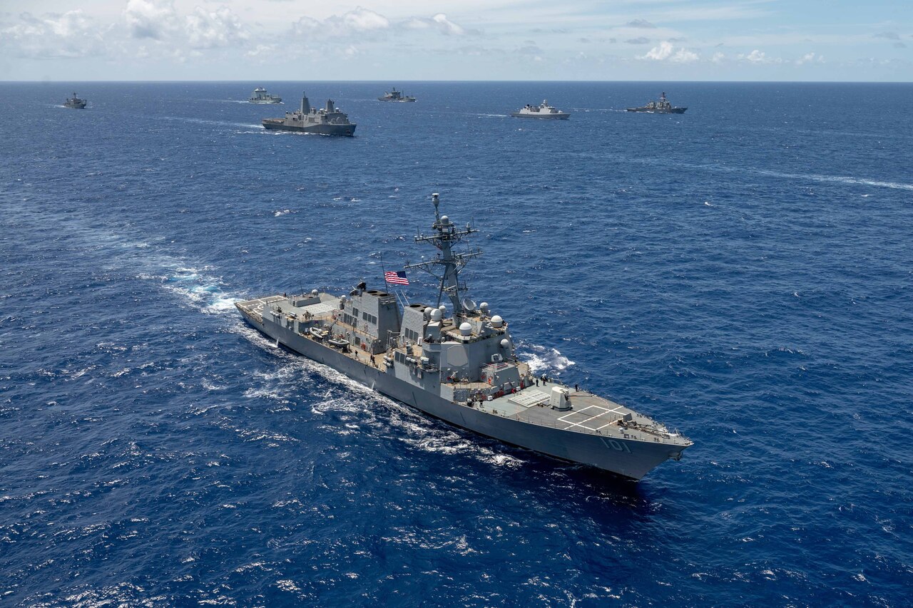 A ship transits a body of water in front of other six other ships sailing in formation during the daylight.