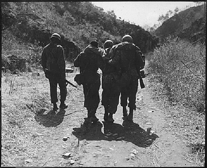 In a black and white photo, two soldiers help a wounded soldier walk along a trail. A fourth soldier walks near them.