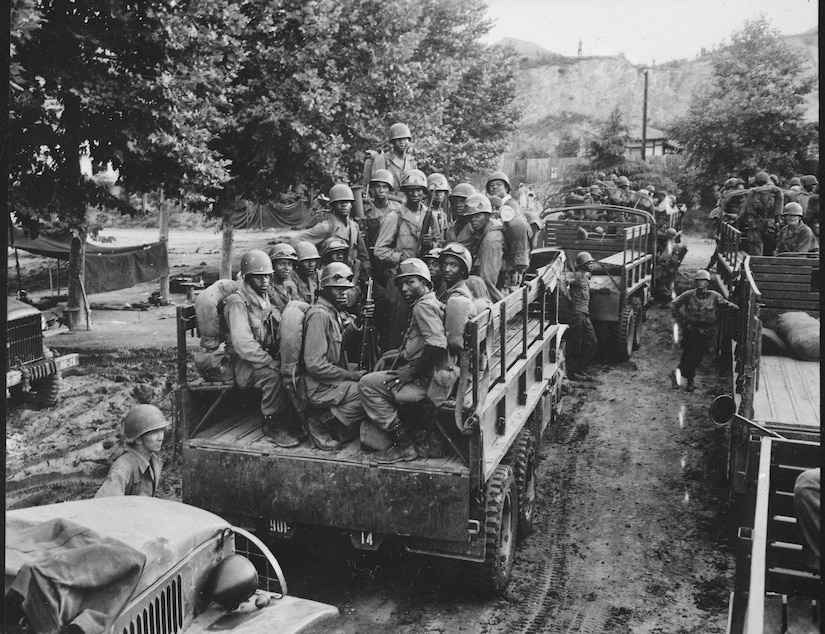 In a black and white photo, soldiers crowd the flatbed of a military vehicle along a road with other vehicles moving in procession.