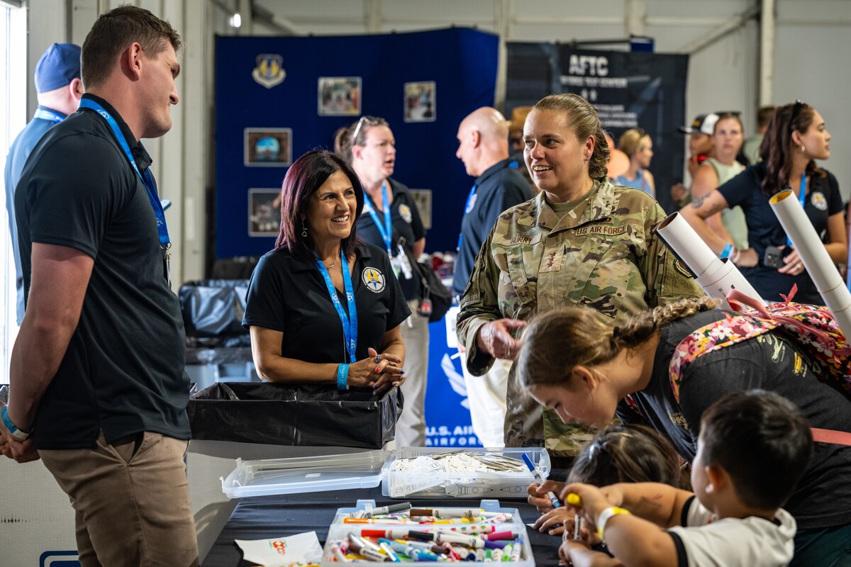 Gen. Hurry smiles as she is briefed by AFMC personnel in black polos, three young attendees are hunched over a table coloring with markers in the foreground
