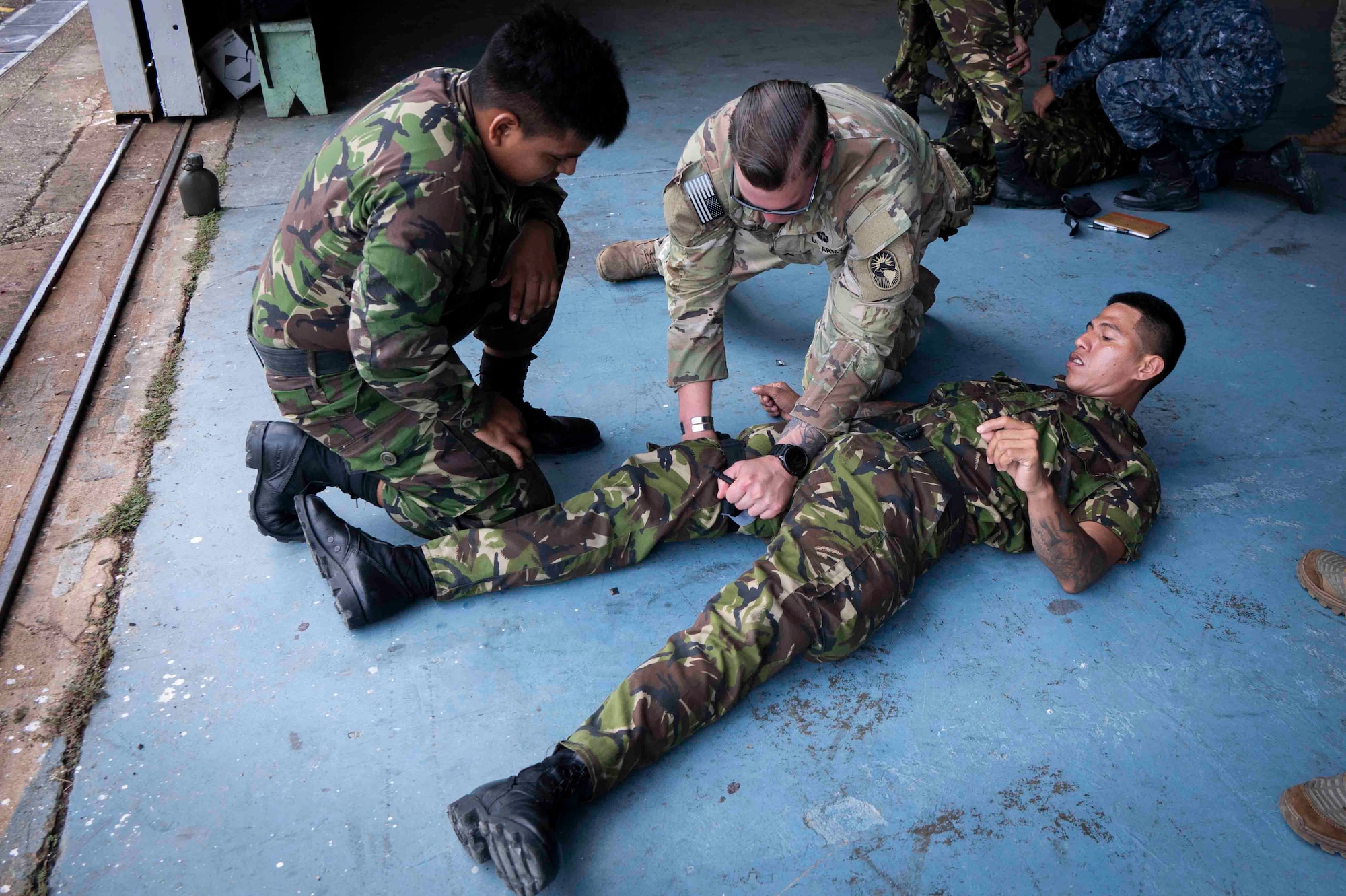 A photo of a U.S. soldier demonstrating how to put a tourniquet on a person's leg while a Belize Solider watches.