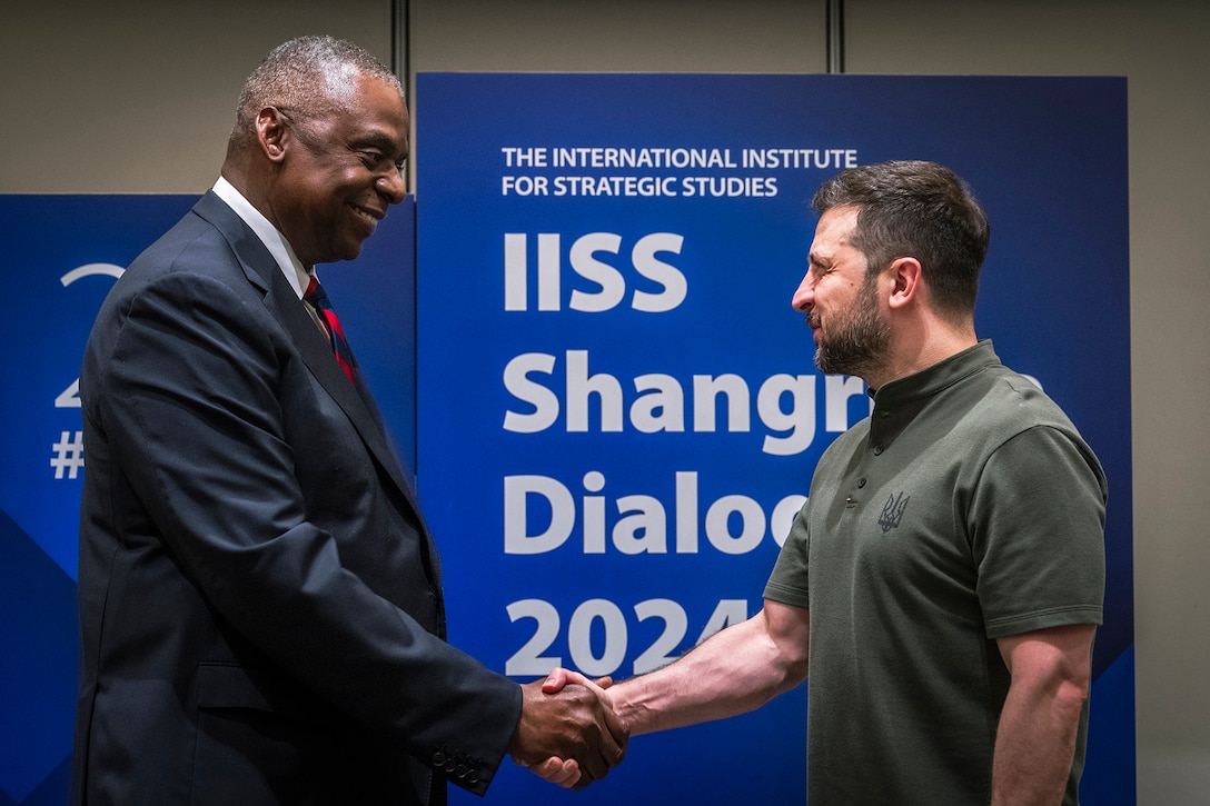 Two people shake hands in front of a blue sign that reads "IISS Shangri-La Dialogue).