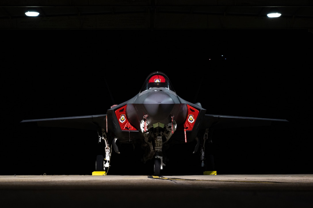A military jet sits under lights on a tarmac at night.
