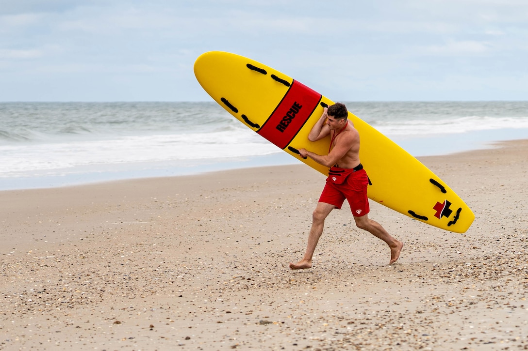 A Marine in swimming shorts runs across a beach carrying a large yellow rescue board.