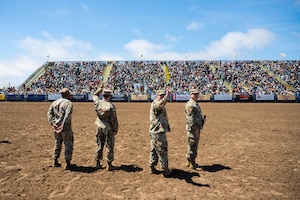 Four U.S. Space Force members wave to a large crowd from the center of a rodeo arena.