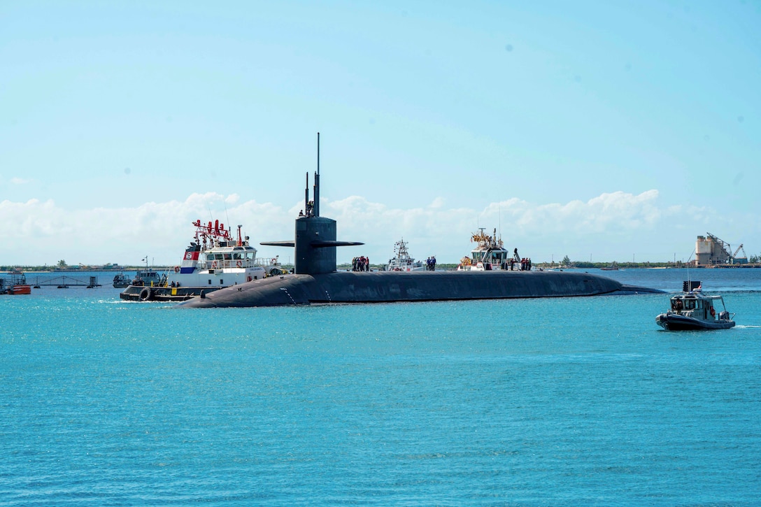 Sailors work on top of a submarine as three boats approach it in a body of water.