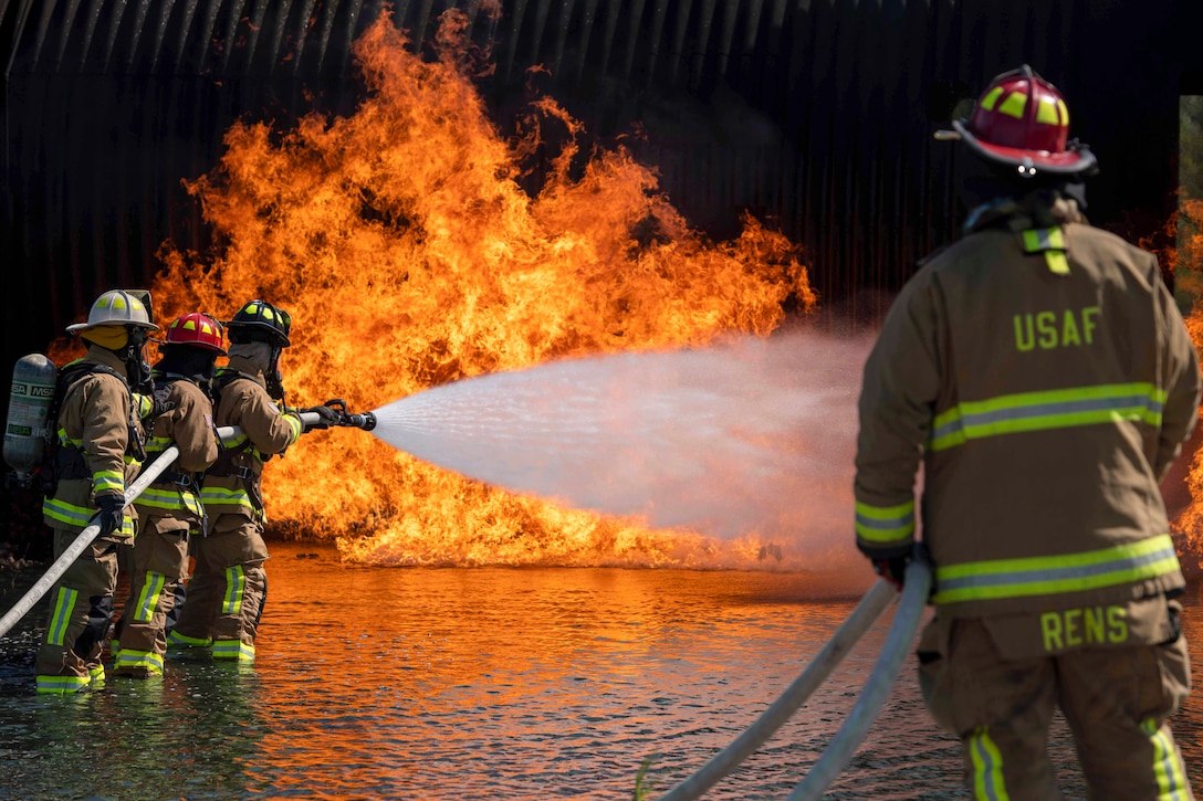 Three firefighters to the left use a hose to put a fire as a fellow firefighter watches from the right as they all stand in a puddle of water.