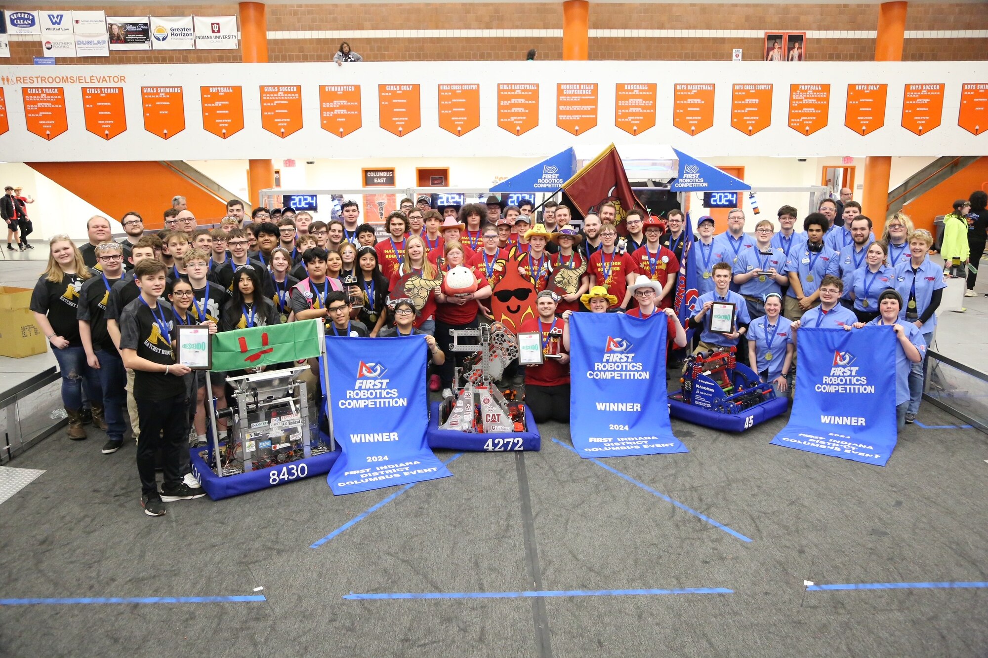 Approximately 80 students pose for a photo. In front of the three distinct groups, each has a robot and a banner front and center.